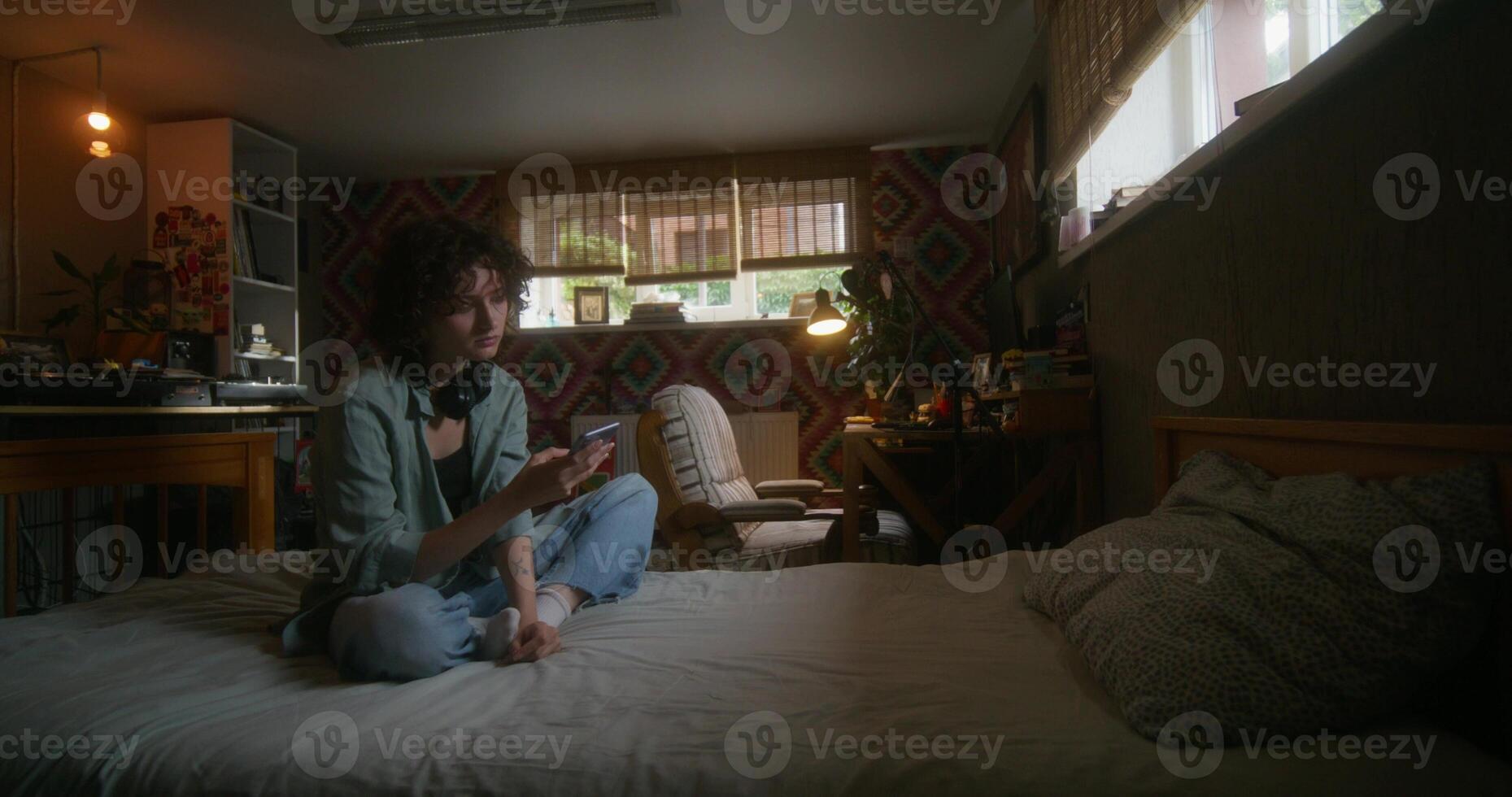 A young woman uses a mobile phone while sitting on a bed at home with a depressed look - sitting - woman photo