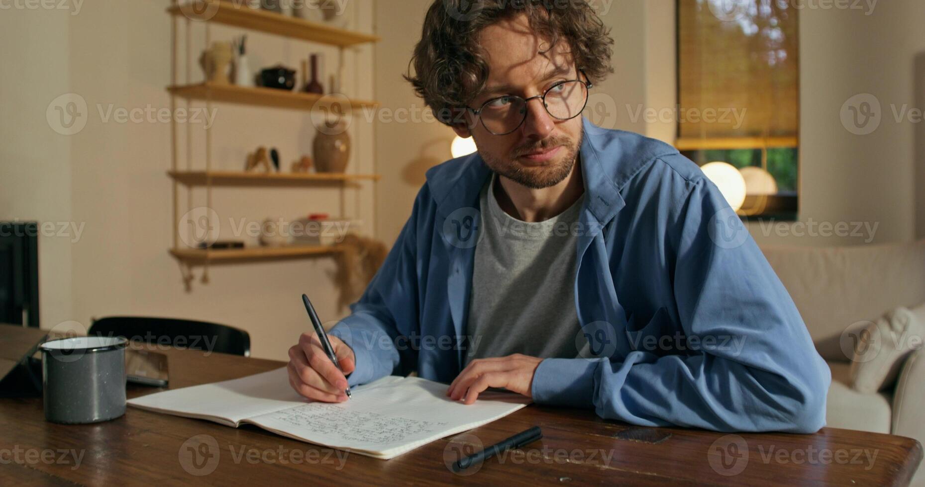 A man with glasses is writing in a notebook sitting at a table with a serious face. Stylish home interior - sitting photo