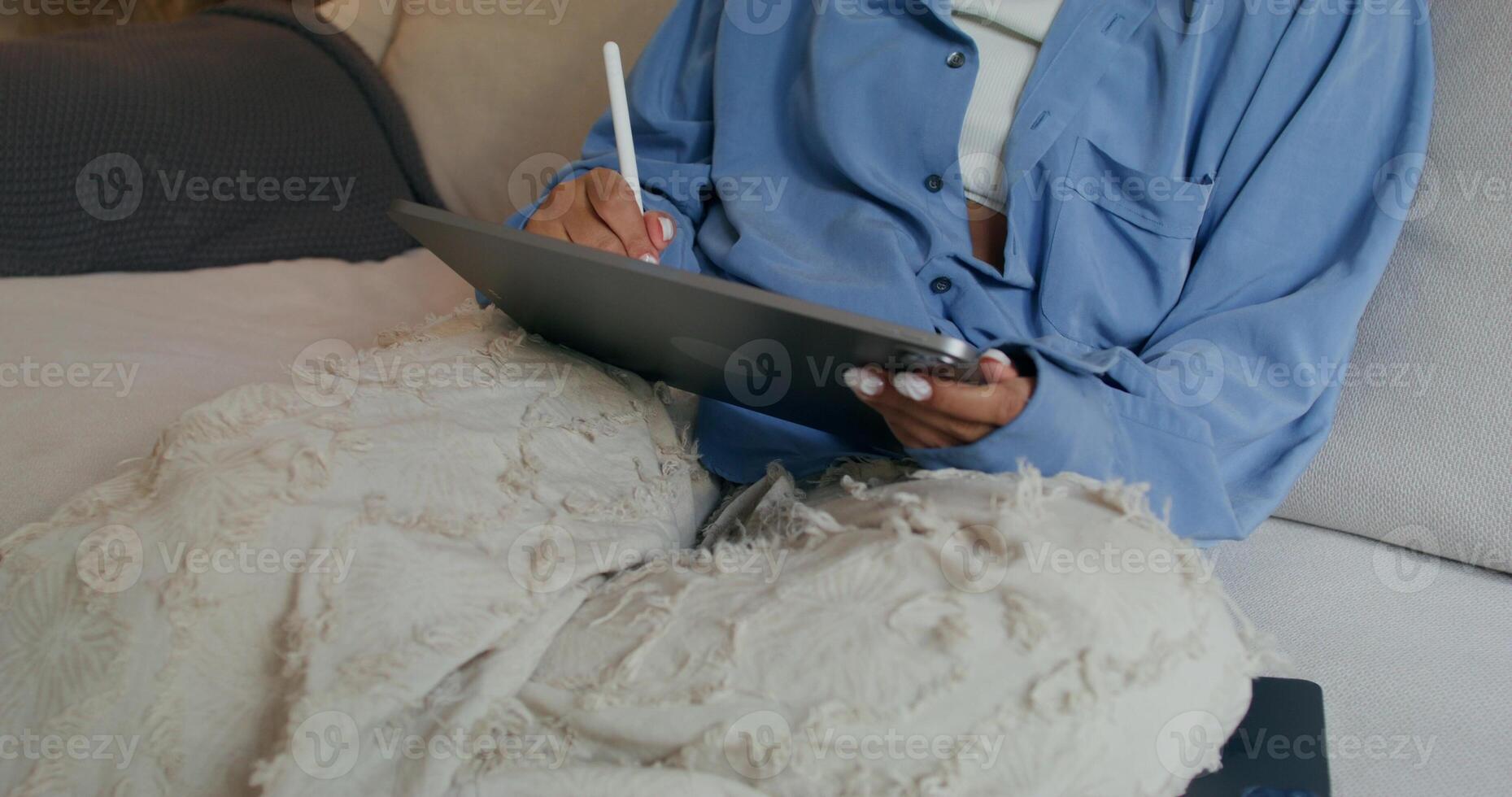 A woman in casual clothes uses a tablet sitting on the couch at home. She draws with a stylus - sitting photo