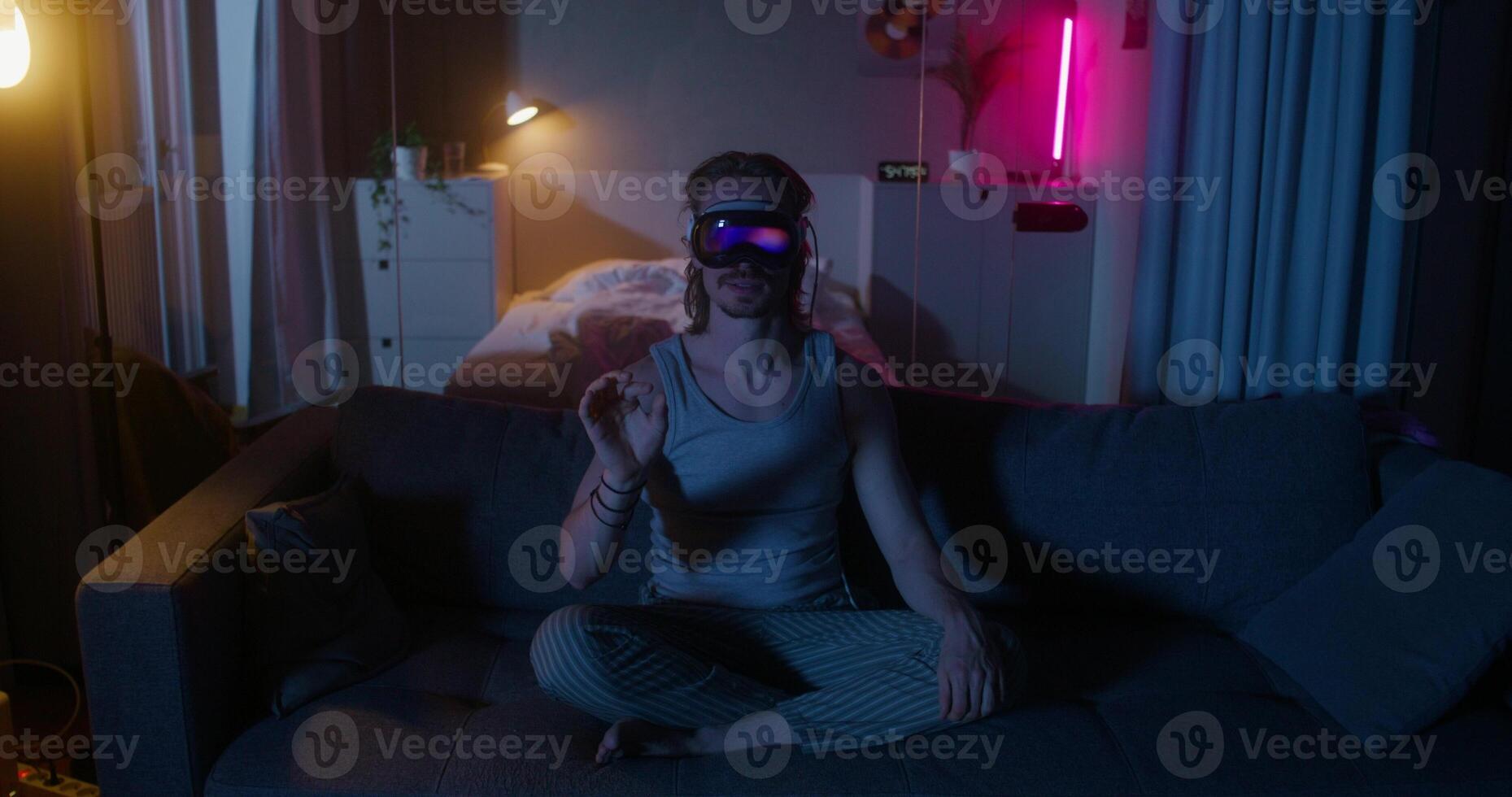 A man wearing virtual reality glasses is sitting on the couch. He's smiling. Dark room, home interior - glasses photo