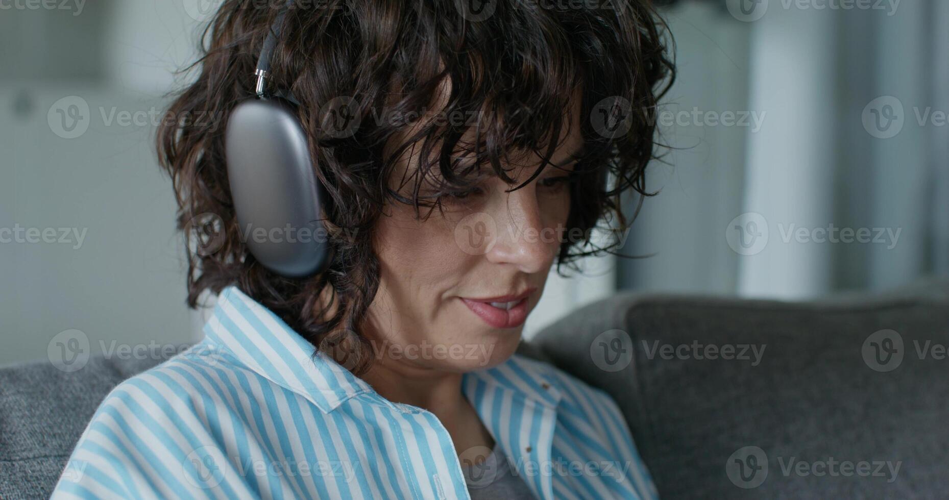 A woman in casual clothes and with headphones is using a mobile phone and smiling. She's sitting on the couch at home. Daylight - Daylight photo