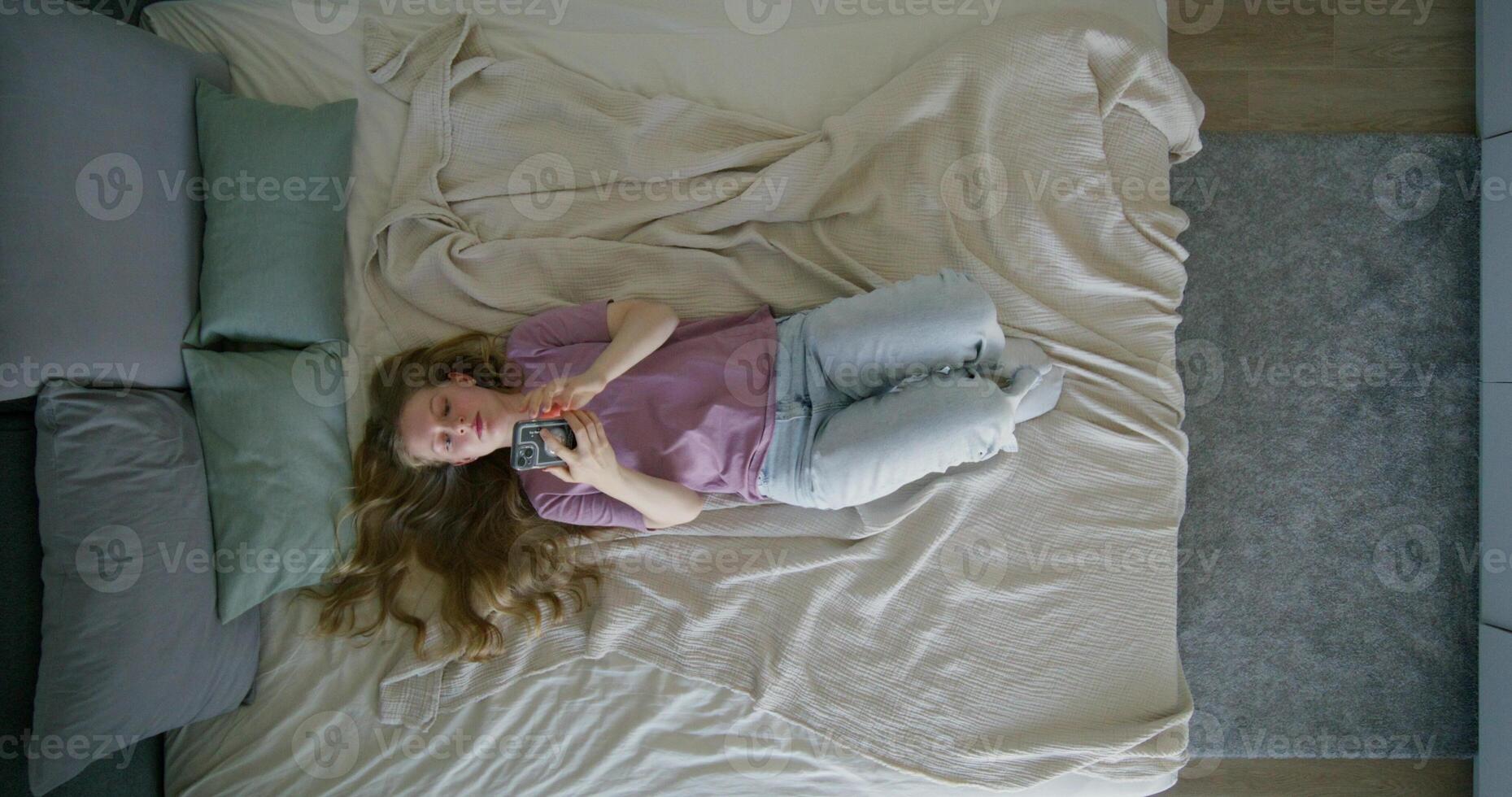 A young woman in casual clothes uses a mobile phone lying on a bed at home. Top view, daylight photo