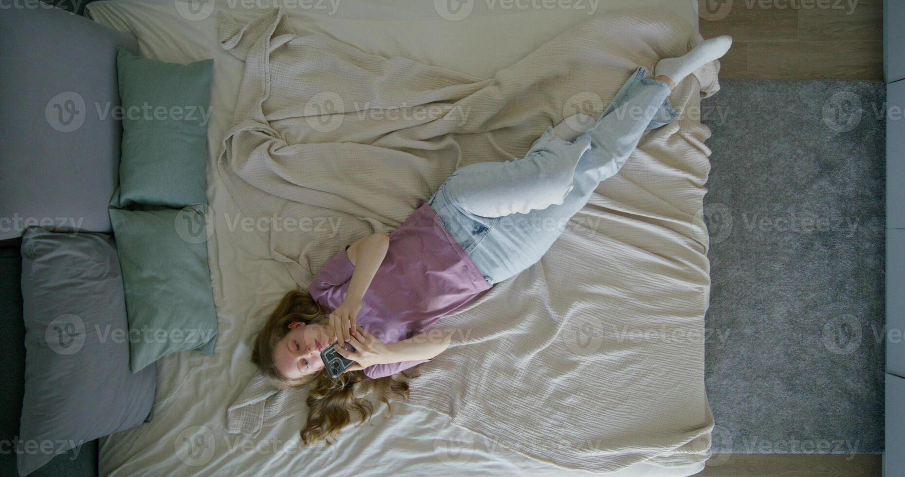 A young woman in casual clothes uses a mobile phone lying on a bed at home. Top view, daylight - daylight photo