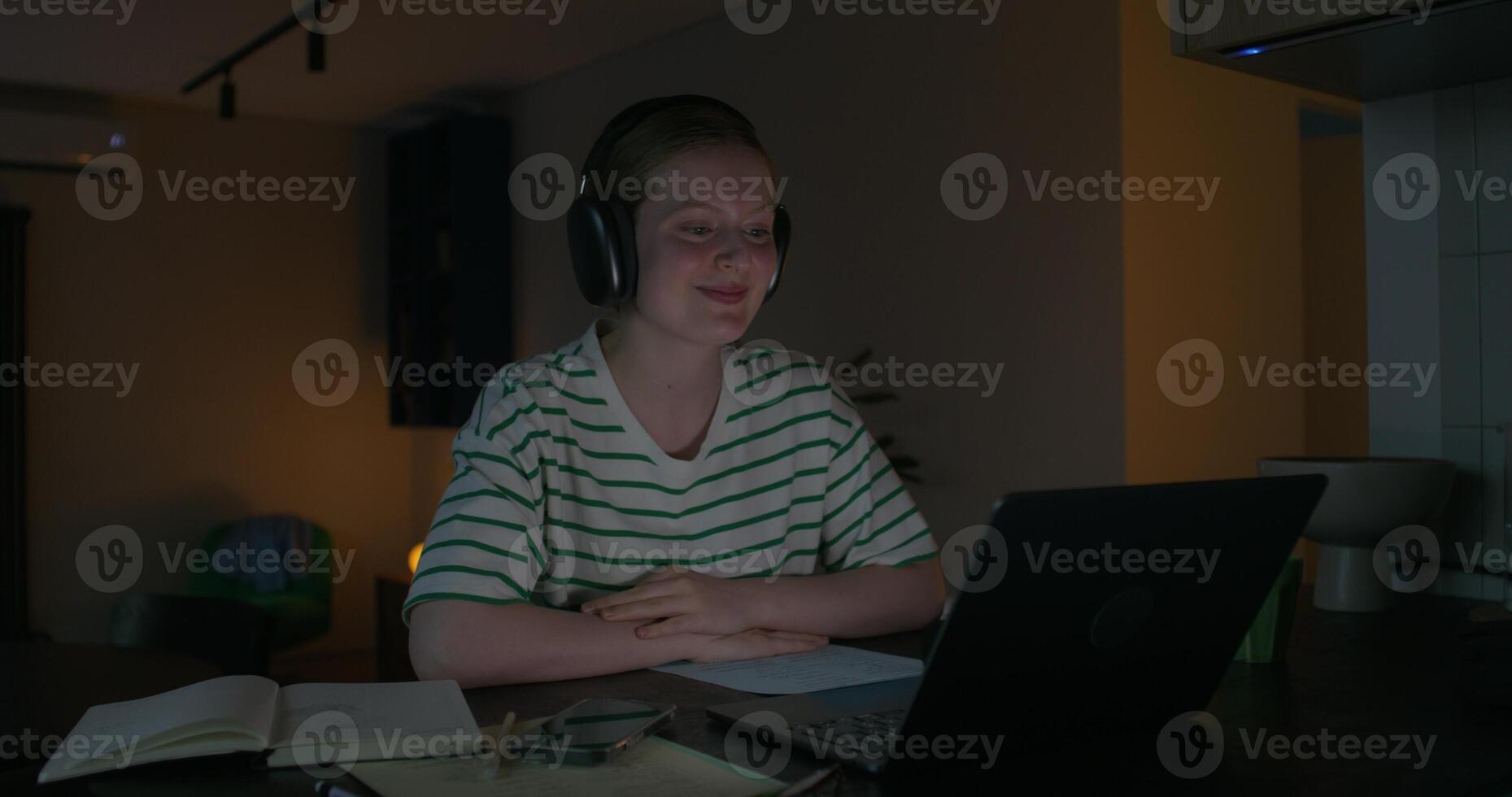 A young woman wearing headphones is smiling and talking on a chat on a laptop. She is sitting at the table at home in the dark photo