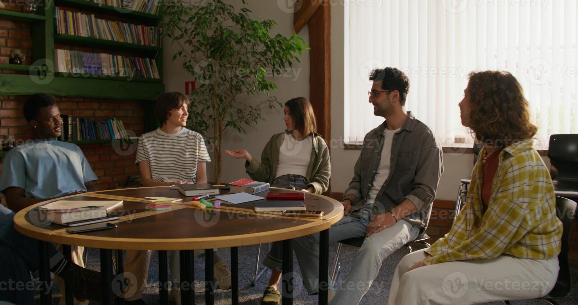 A group of young people are smiling and talking while sitting around a round table indoors. Notebooks and books on the table. Daylight - talking photo