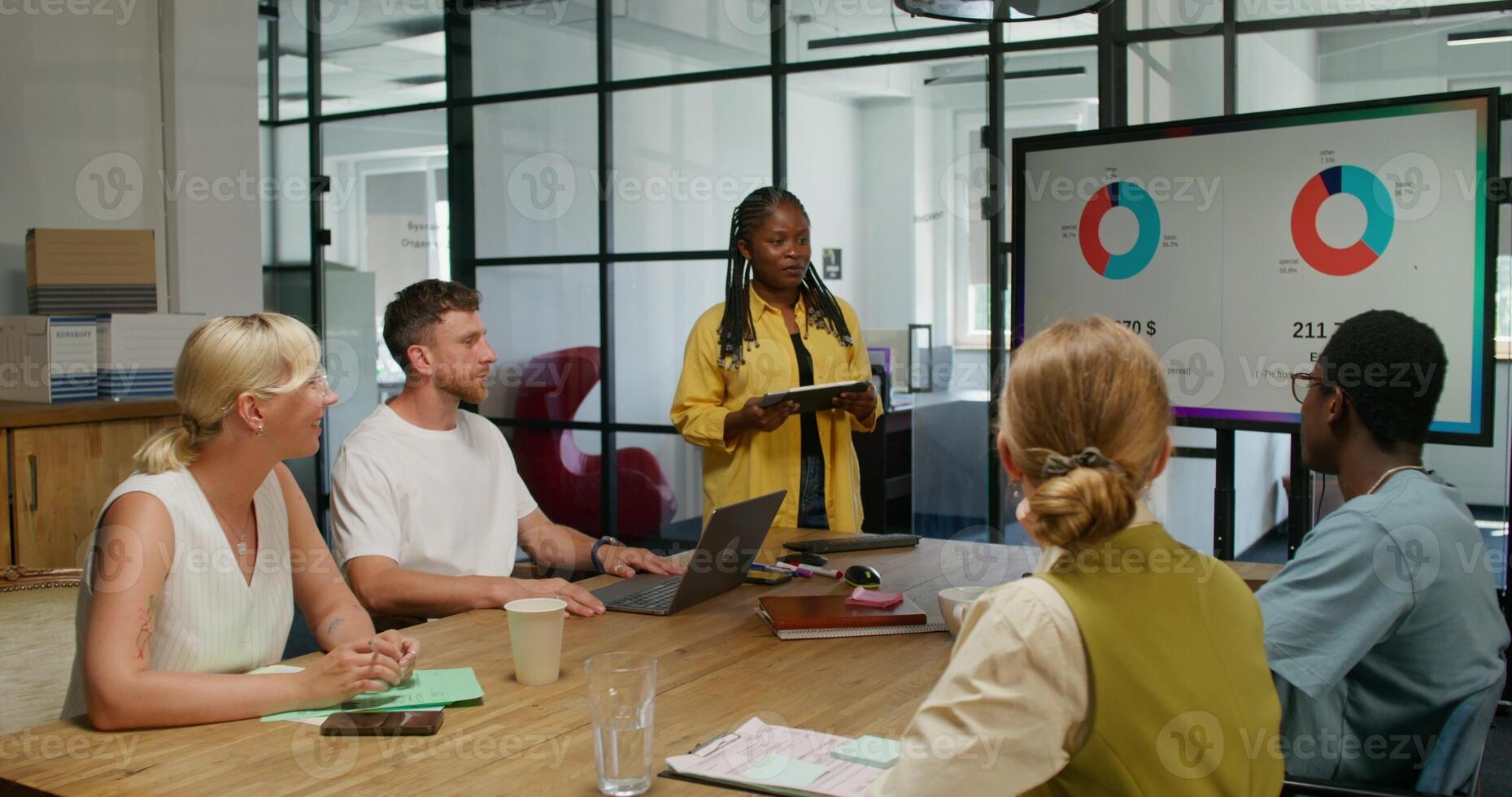A group of young people are sitting at a table in an office meeting. A female speaker is talking standing near the screen. An open laptop is in front of a man. Daylight photo