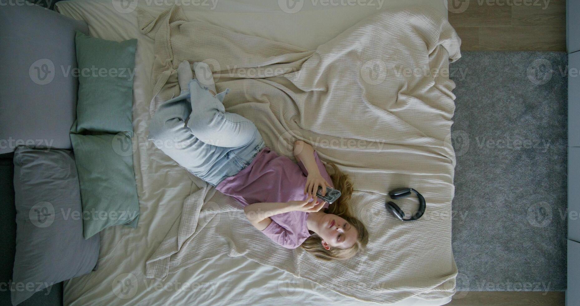 A young woman in casual clothes uses a mobile phone lying on a bed at home. Top view, daylight - mobile photo