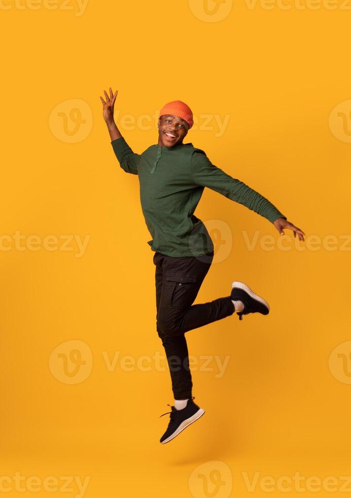 Cheerful Excited Black Hipster Guy Jumping In Air Over Yellow Background, Millennial African American Man In Orange Hat Having Fun In Studio, Looking And Smiling At Camera, Full-Length, Copy Space photo
