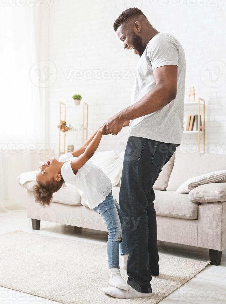 Black dad teaching little naughty daughter dancing at home, girl staying on father feet and having fun photo