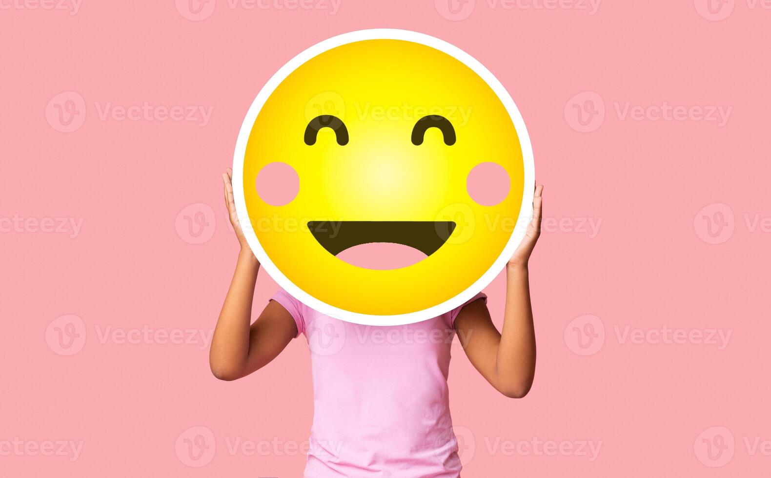 Black girl hiding face behind happy shy emoji emoticon, standing over pink studio background with copy space photo