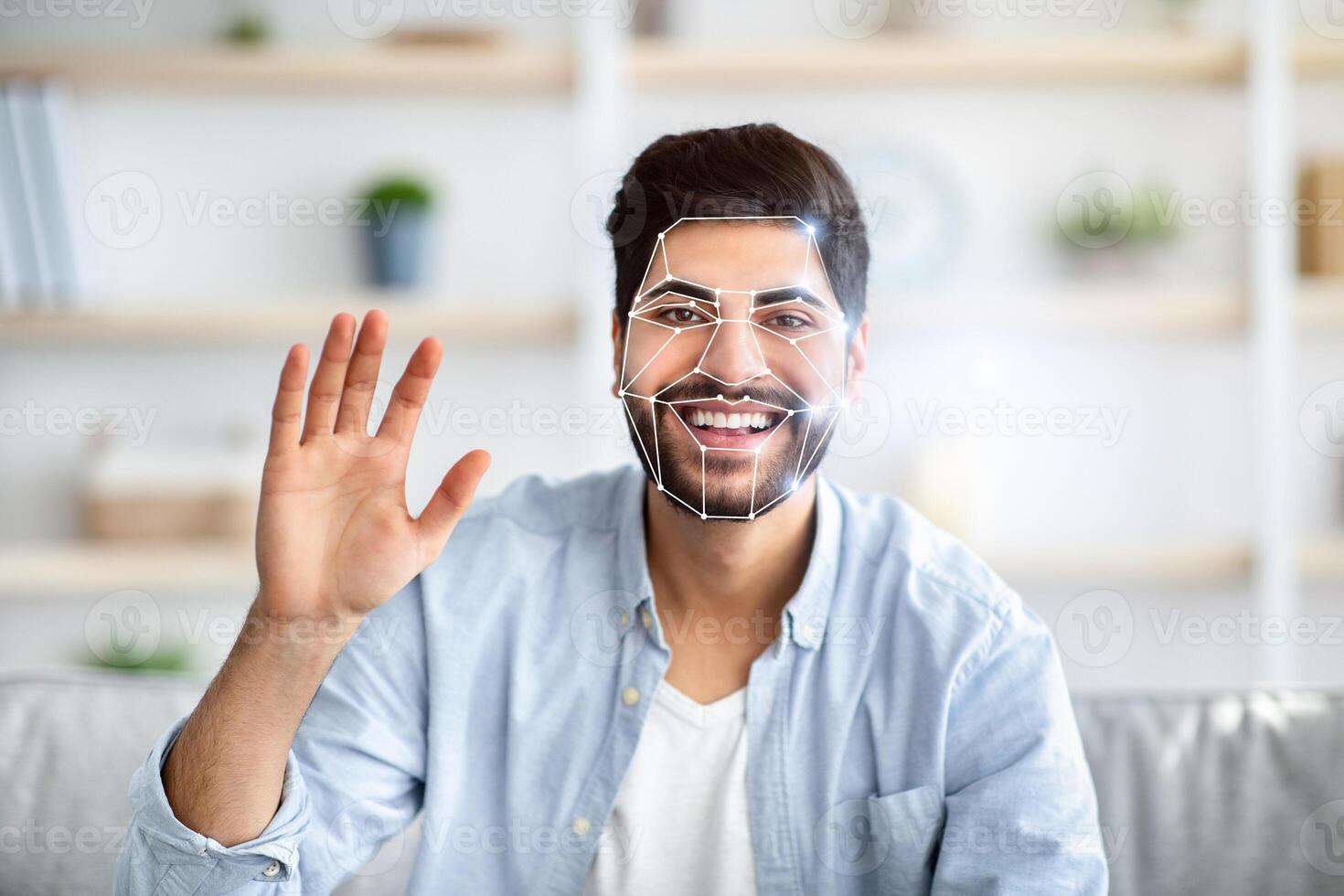Indian man is using facial recognition technology in a home setting. He is smiling while waving, with a digital overlay highlighting his facial features for identification. photo