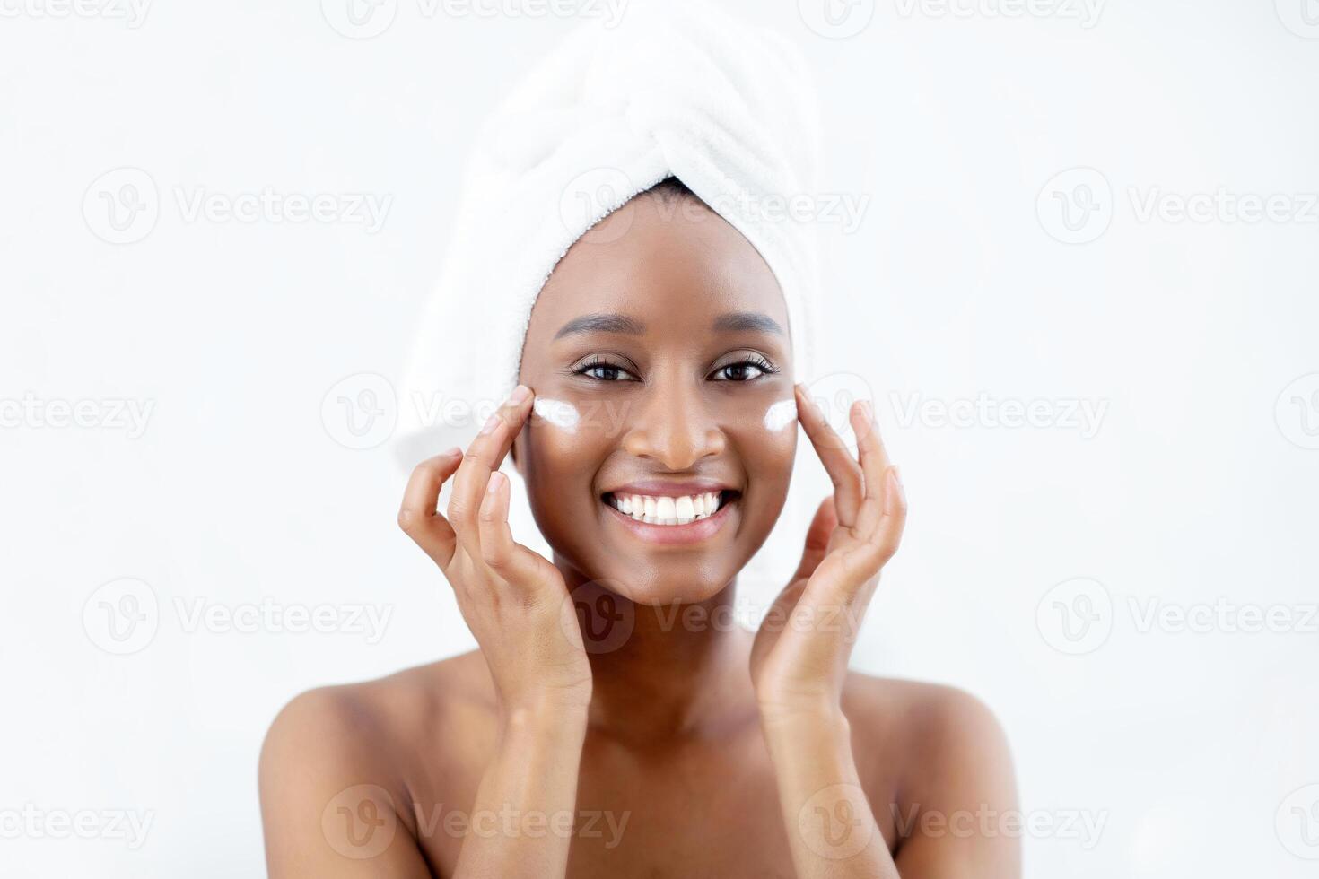 Skin hydration, anti-aging care and home spa. Cheerful attractive millennial african american woman with towel on head after shower applies cream on face, in bedroom in morning, on white background photo