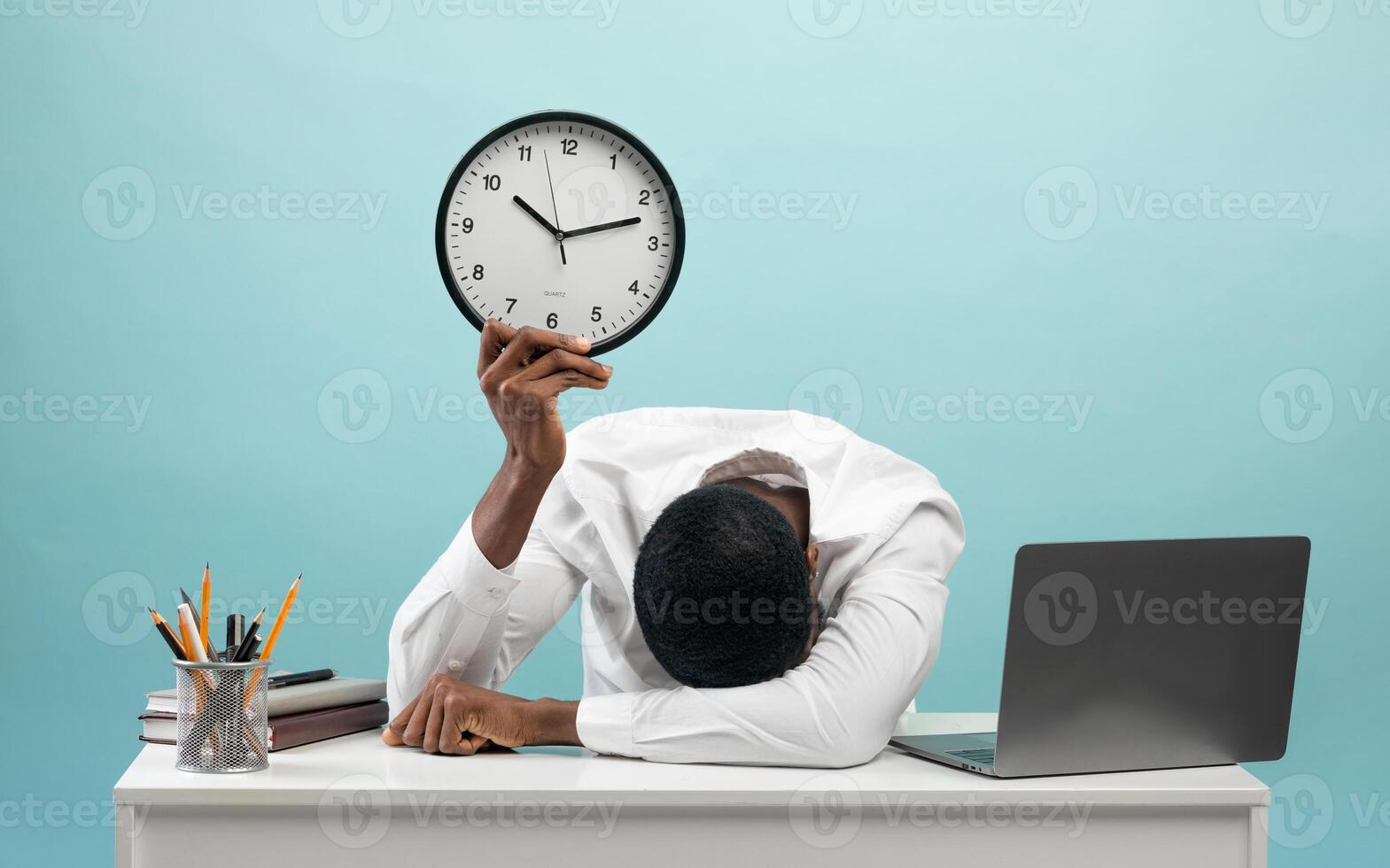 Tired african american businessman sitting at desk with pc laptop, holding clock and putting head on hand, feeling exhausted over blue studio background photo