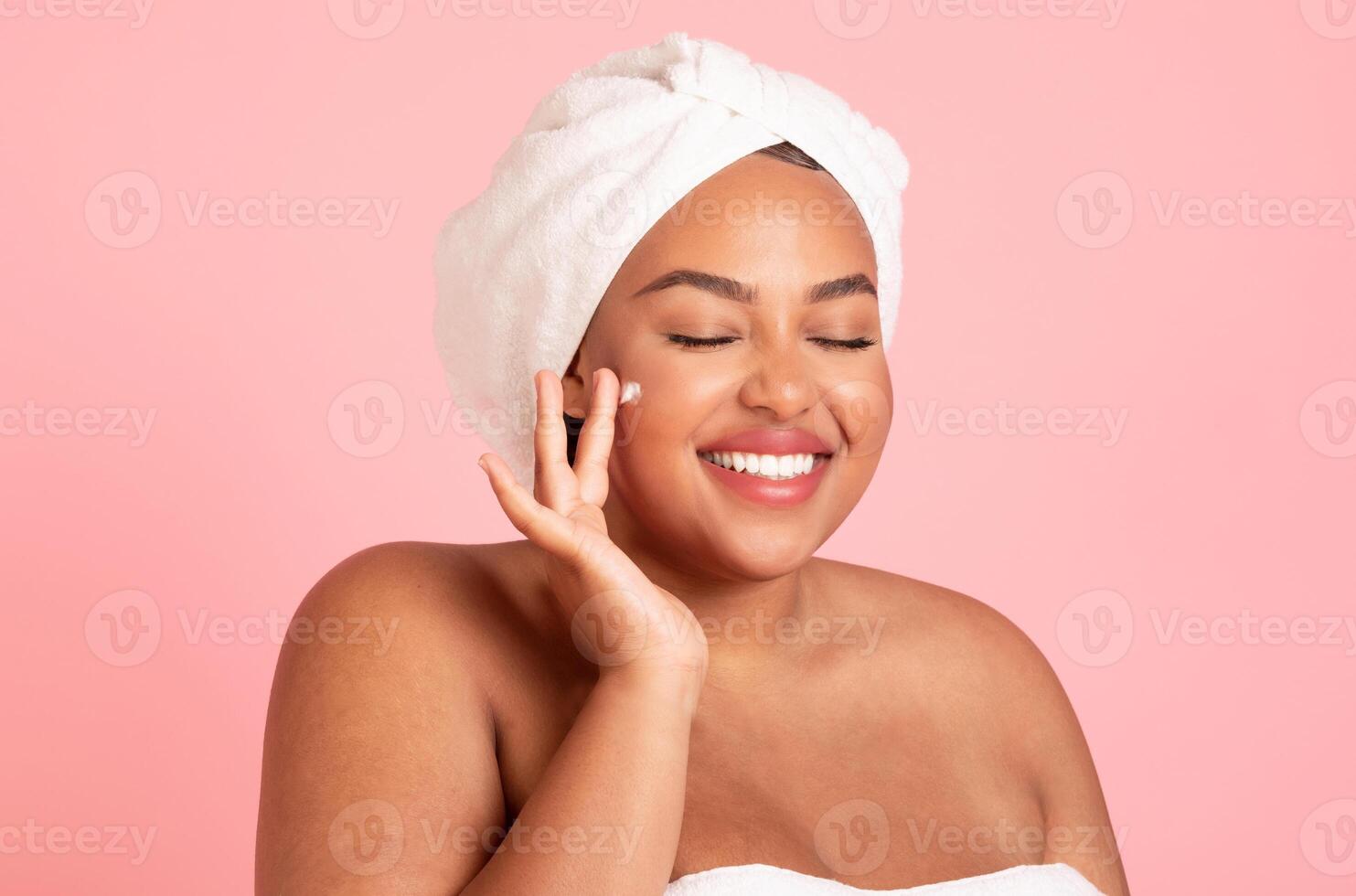 Happy black body positive woman applying beauty product on her face and smiling, lady wrapped in towel using face cream after shower, isolated on pink studio background photo