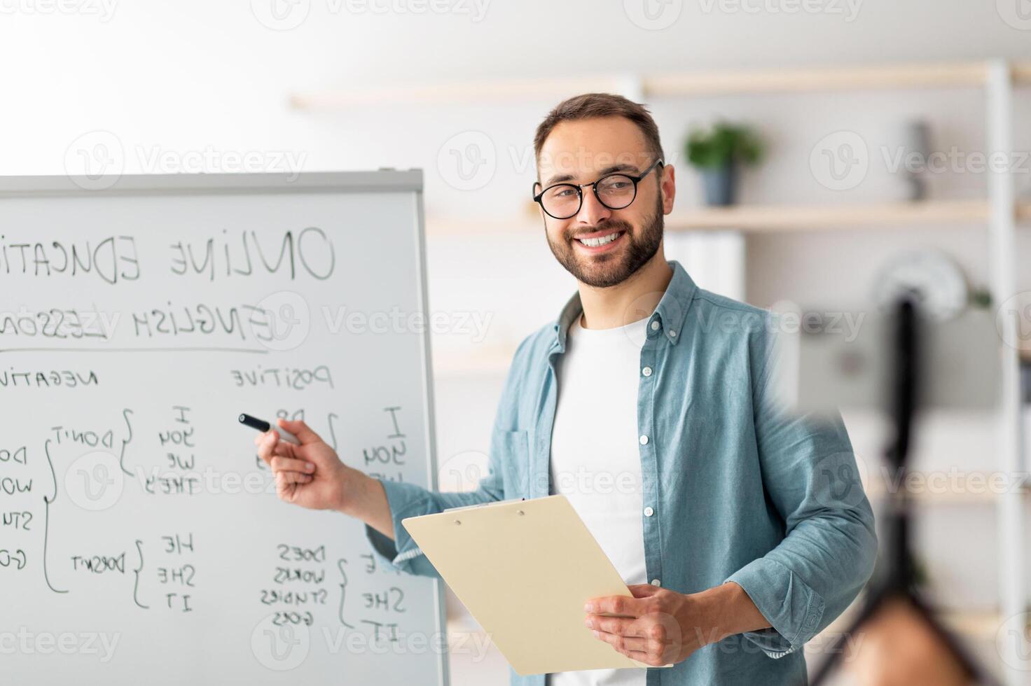Portrait of happy young male teacher standing near blackboard with English grammar rules, conducting internet lesson. Positive tutor giving oreign language class on call or web conference photo