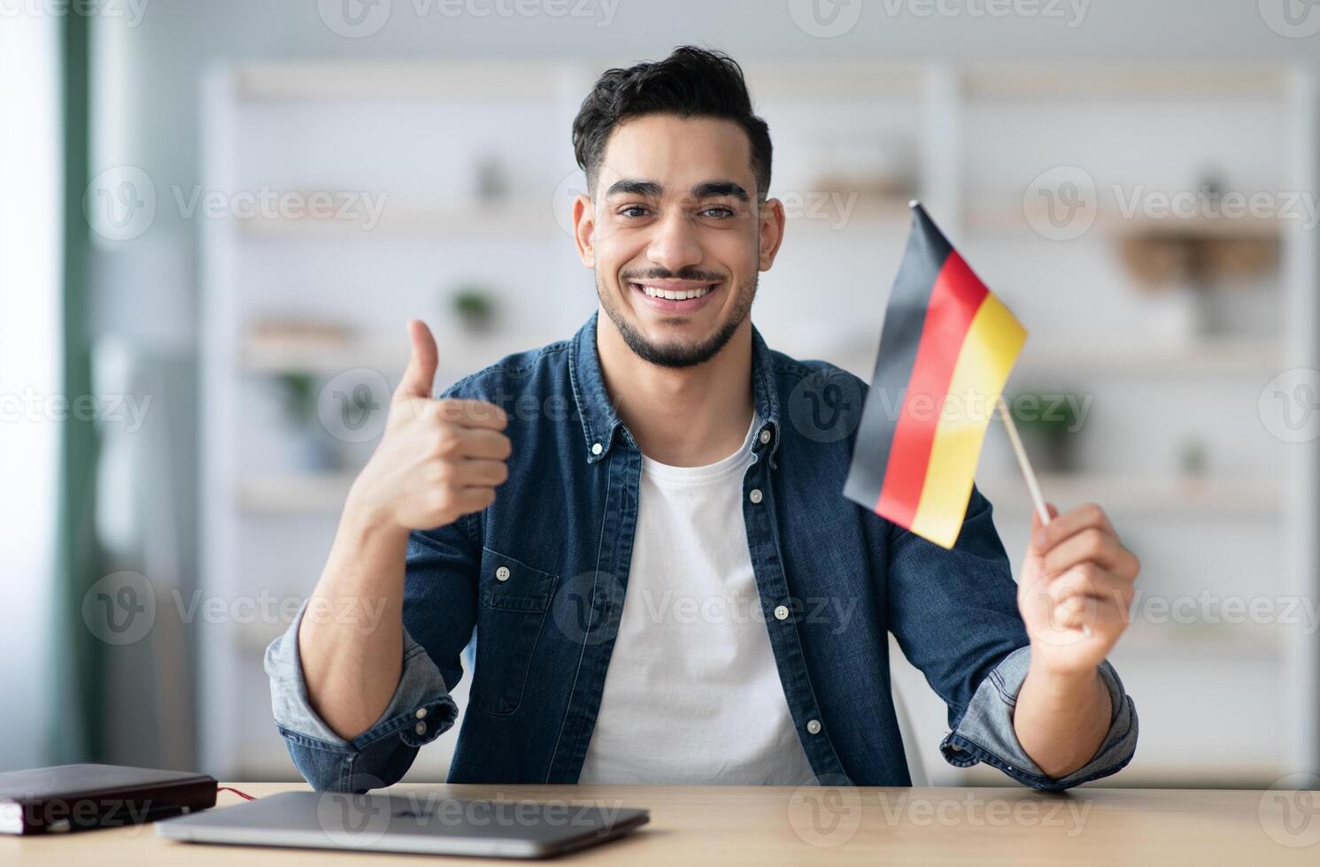 Smiling arab guy with flag of Germany showing thumb up, sitting at desk with laptop, middle-eastern young man student learning German for job, education or emmigration to Germany, copy space photo