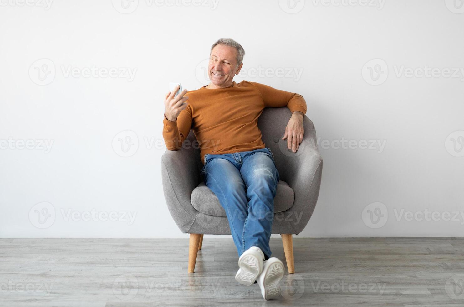 Portrait of smiling mature man using mobile phone, watching sitting resting on armchair isolated at white background wall. Guy browsing internet, surfing web, free copy space, full body length photo