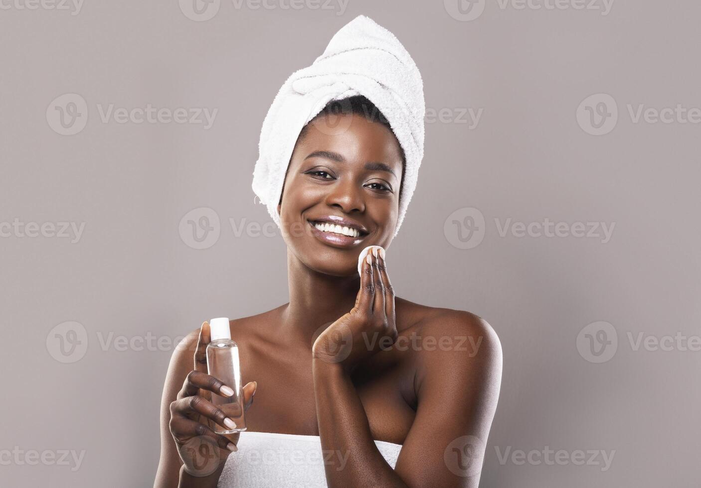 Young beautiful african woman cleaning her face from makeup with cotton pads and micellar water over gray background with copy space photo