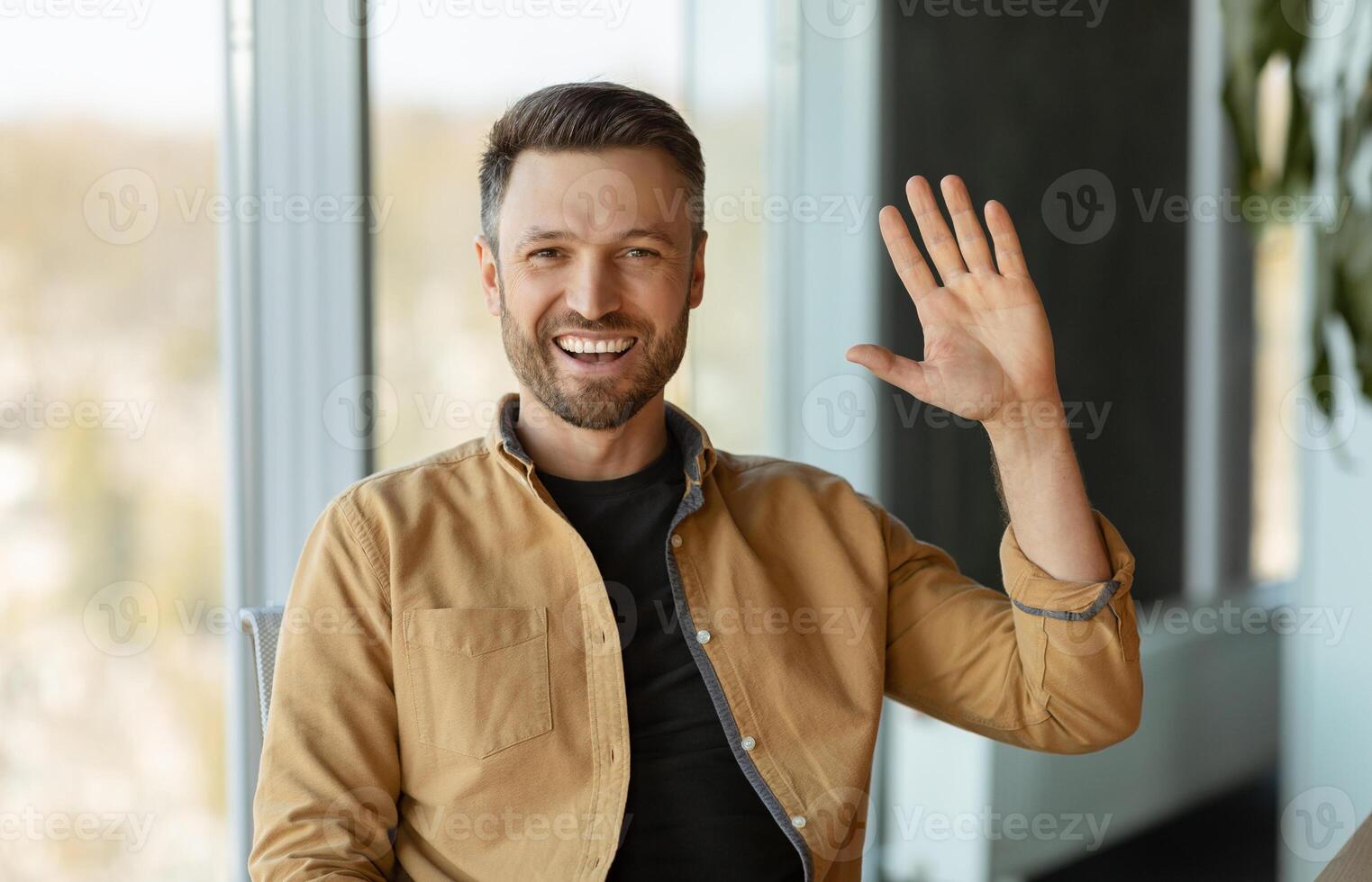 Joyful Businessman Waving Hello Smiling To Camera Sitting In Modern Office. Entrepreneur Man Communicating Online Gesturing Hi Greeting At Workplace. Business Communication Employment photo