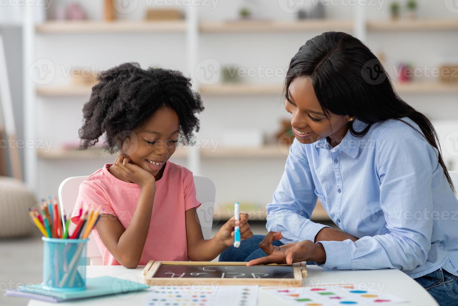 Friendly young african american woman speech therapist teaching adorable little kid alphabet, drawing letters on blackboard with chalk. Speech-language therapy for children concept photo