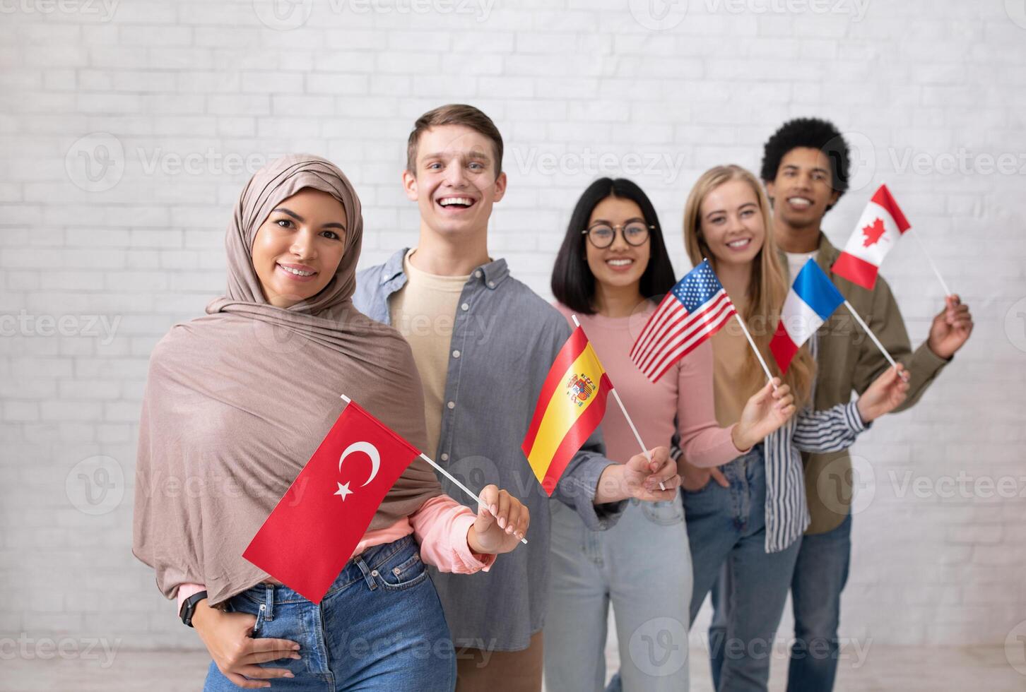 Modern exchange program and language learning. Happy young international students looking at camera and holding flags of their countries, on white brick wall background, studio shot, copy space photo