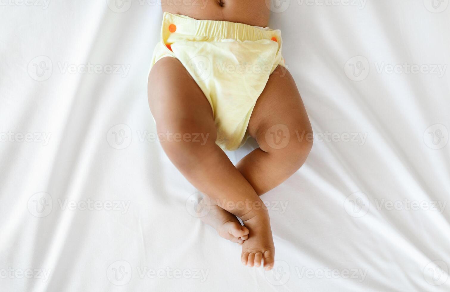 Bottom Part Of Black Infant Baby Wearing Reusable Diaper Lying On Bed, Top View Of Unrecognizable Little African American Boy Or Girl Relaxing On White Linens In Bedroom, Cropped Shot With Copy Space photo