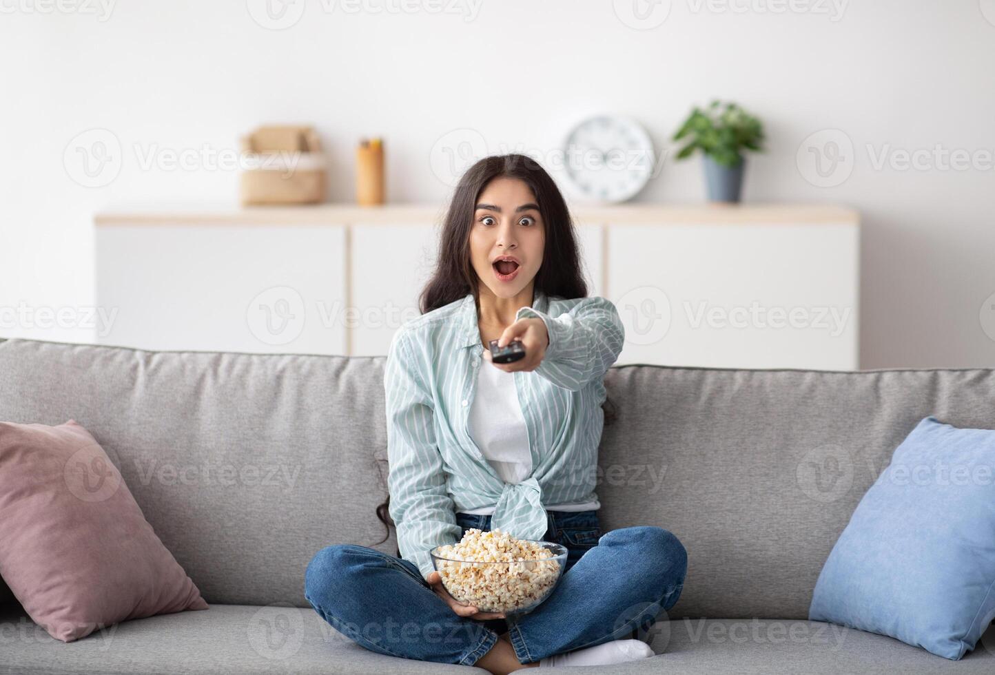 Full length of shocked Indian woman sitting on sofa, eating popcorn, switching channels, watching TV at home. Surprised young lady with remote control enjoying interesting movie indoors photo