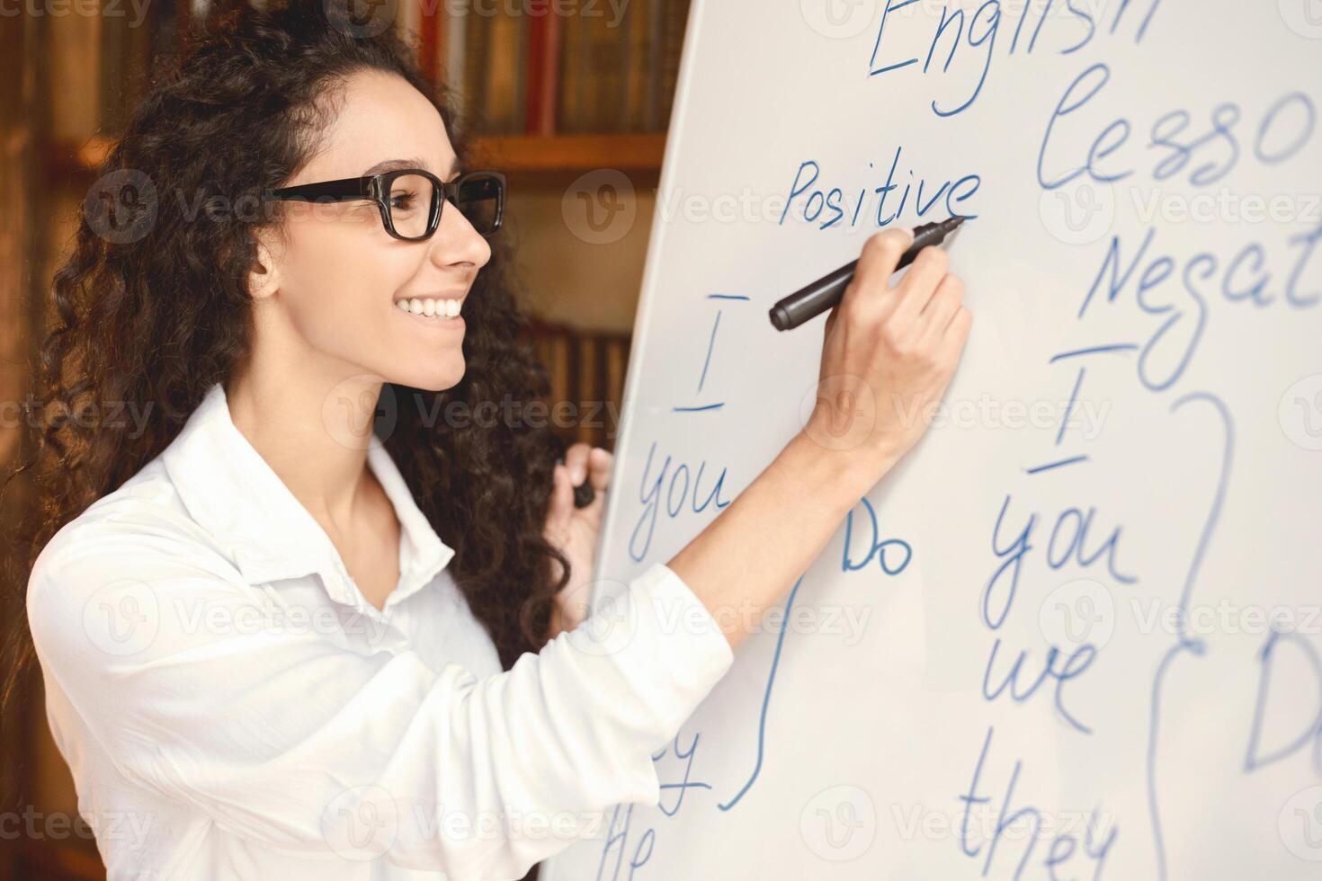 Education Cocnept. Smiling professional English teacher standing at whiteboard, writing with marker. Young female lecturer wearing glasses, looking at board, explaining grammar rules to the students photo