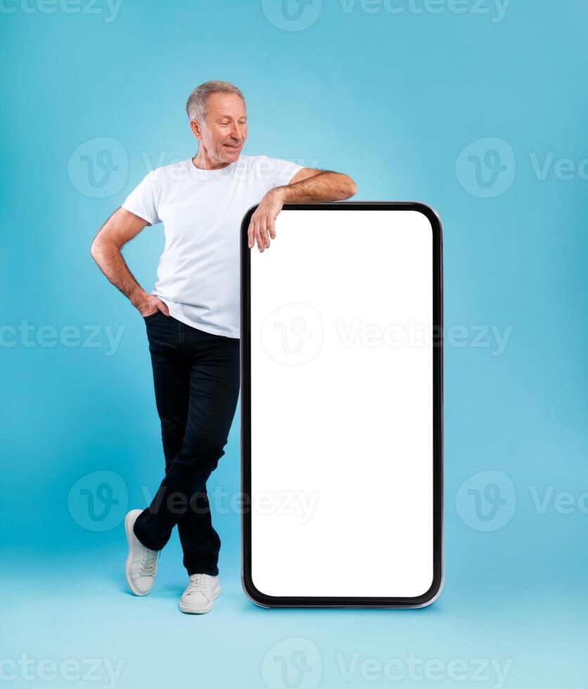 Cheerful smiling mature man looking at giant big cell phone, leaning on empty white screen template panel, standing on blue studio wall. Mock up for app or website, free copy space, full body length photo