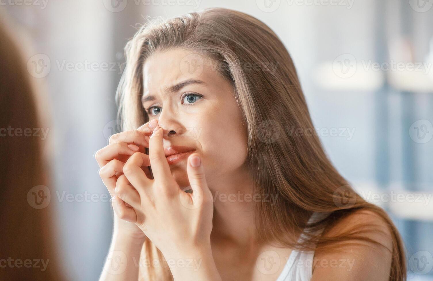 Displeased young woman squeezing acne on her nose while looking in mirror at home photo