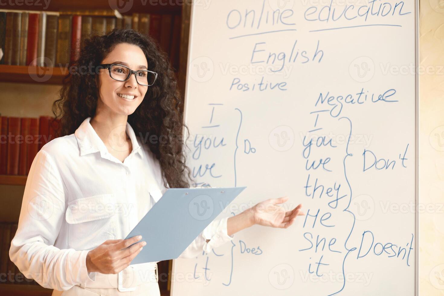 Lecture And Online Education Concept. Portrait of smiling positive young lecturer wearing eyeglasses teaching English language, pointing at grammar rules on board, holding clipboard, explaining photo