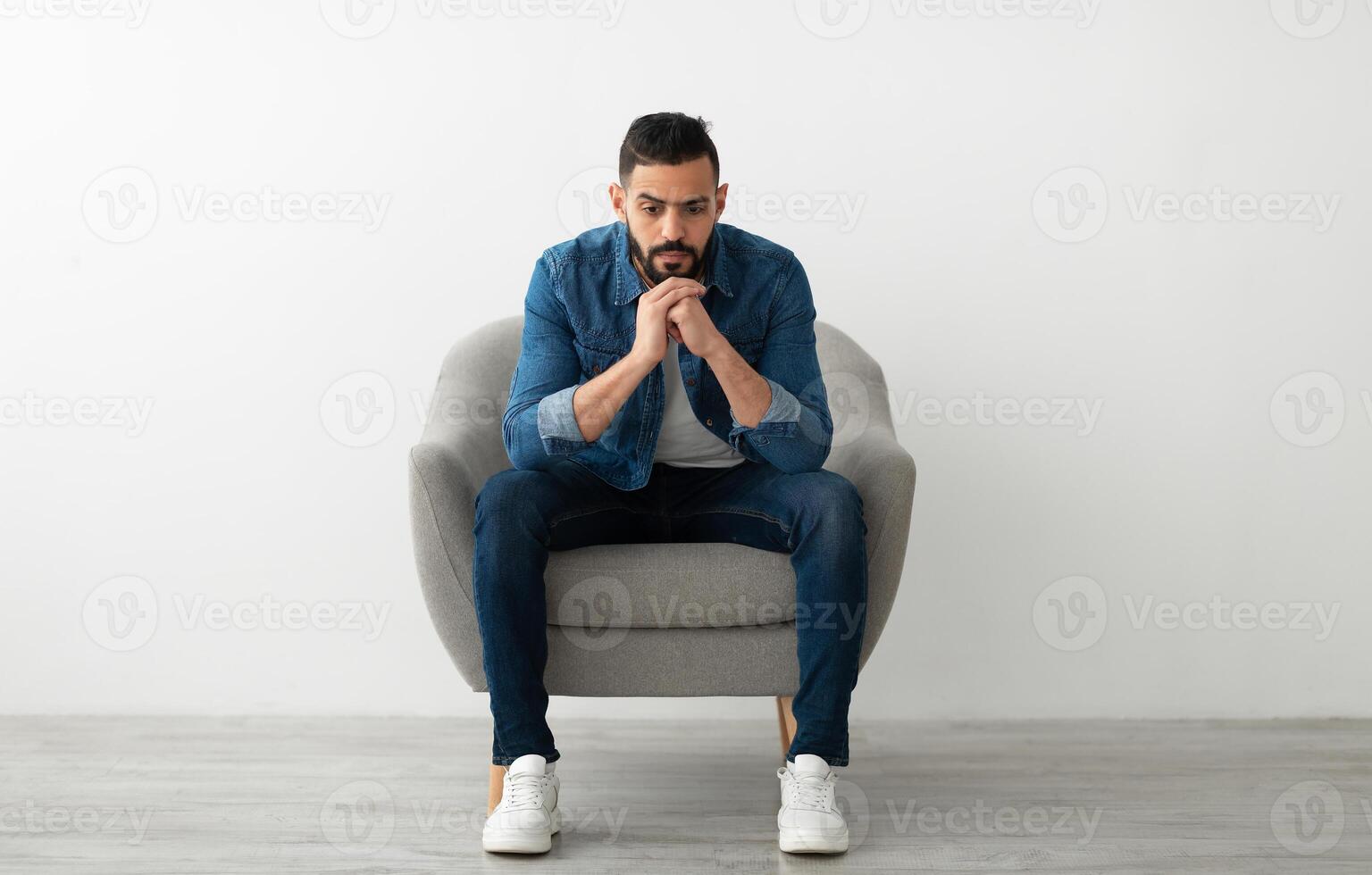 Negative emotions. Young Arab man sitting in armchair with dull or thoughtful face expression, feeling unhappy or tired, having problem, suffering from depression against white studio wall photo
