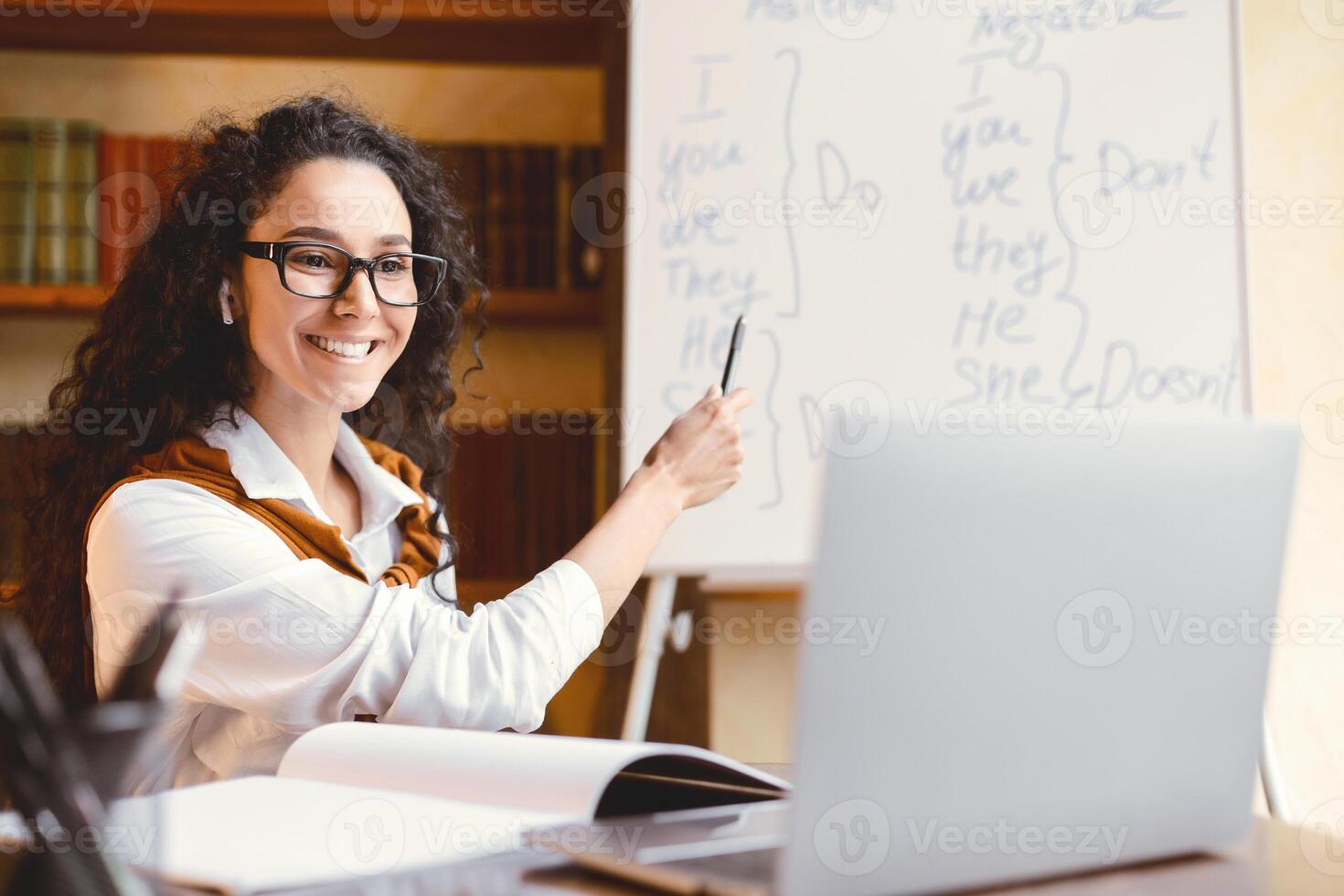 Education And E-Learning Concept. Portrait of smiling female English teacher sitting at table, pointing with pen at whiteboard, explaining grammar rules. Excited lady wearing glasses and earphones photo