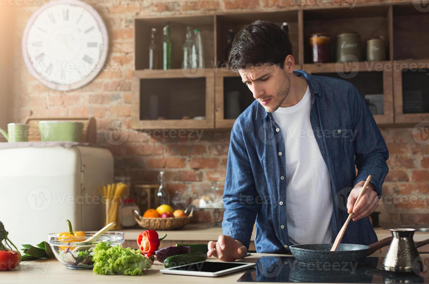 Happy man cooking healthy food with recipe, looking on digital tablet in loft kitchen at home on sunny day. Preparing vegetable salad. photo