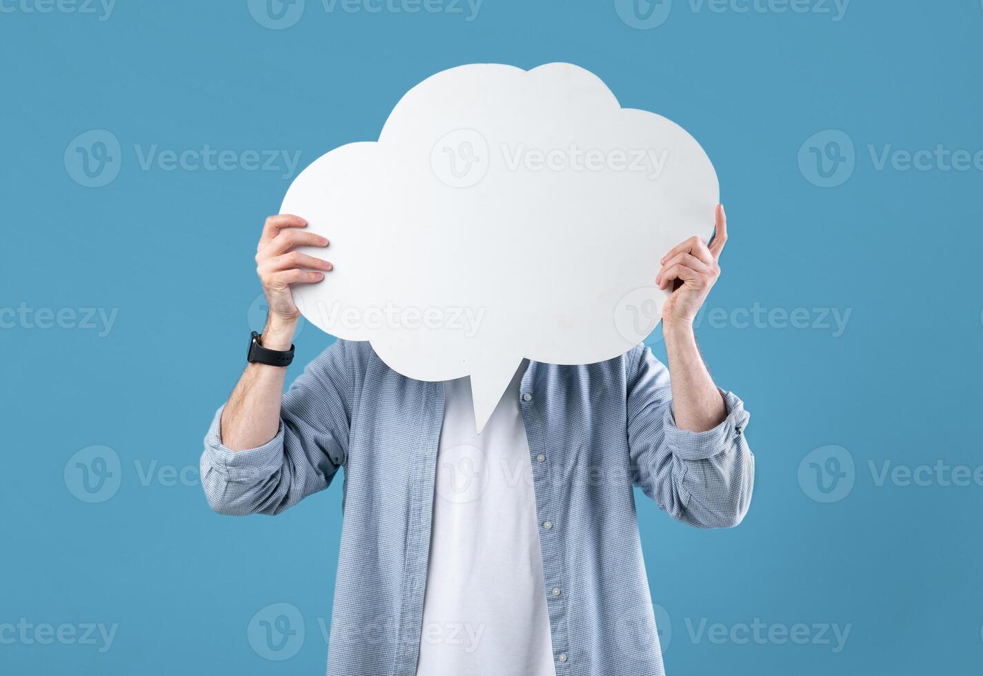 Faceless young man covering his face with speech bubble on blue studio background, mockup for design. Anonymous millennial guy hiding behind word cloud, space for your ad photo