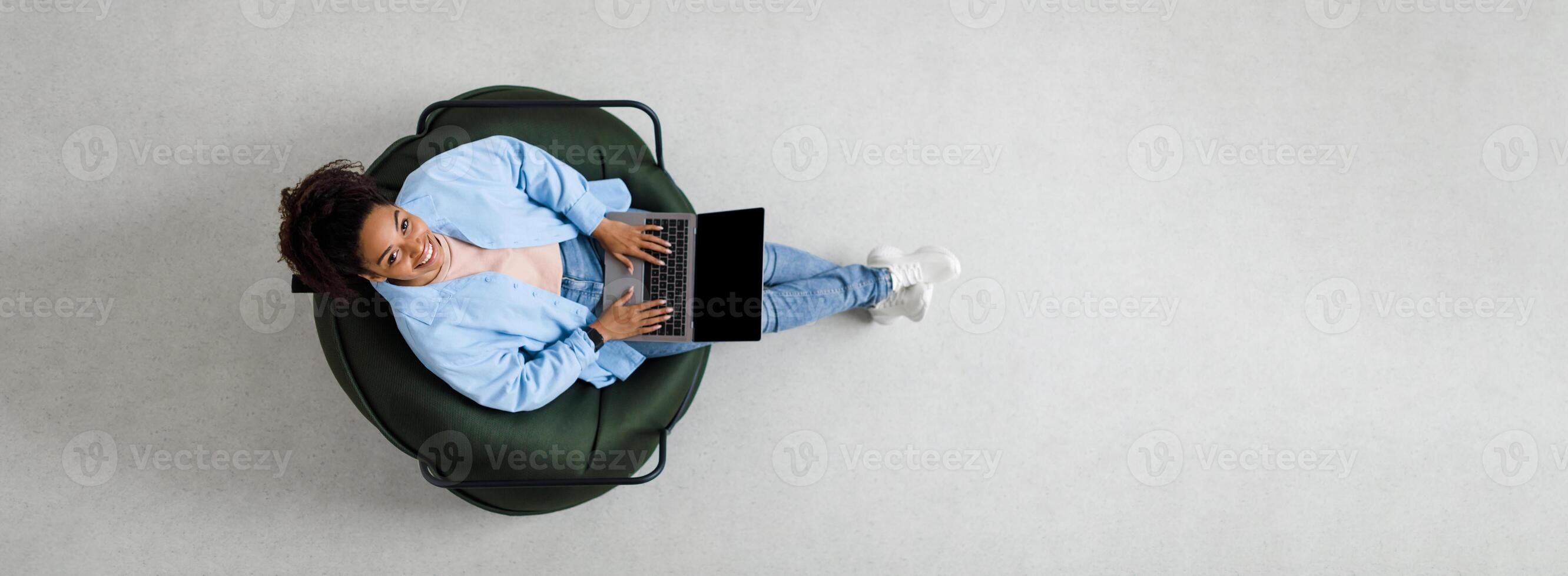 People And Modern Technology. Above top view of young smiling black woman holding pc on lap sitting on bean bag in living room, looking up at camera. Lady browsing internet, free copy space panorama photo