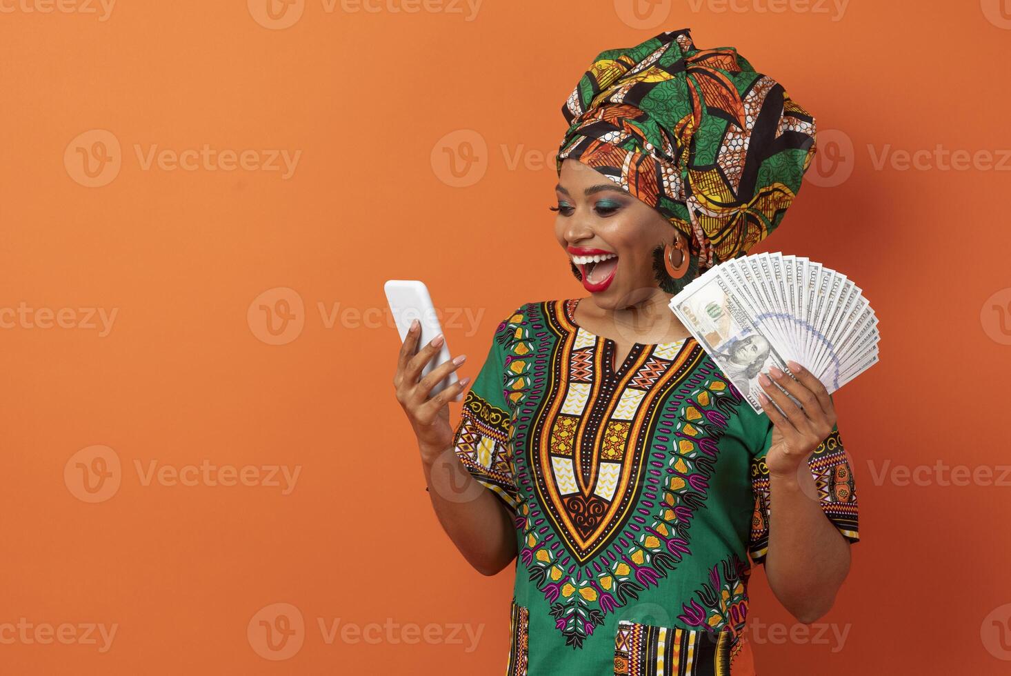 Emotional happy millennial african american lady in traditional costume with money cash dollar banknotes and smartphone in her hands exclaiming, isolated on orange background, copy space photo
