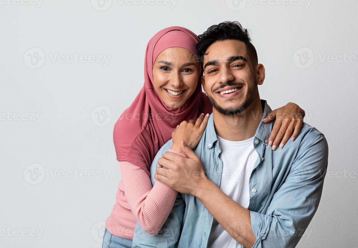 Portrait of romantic smiling muslim couple posing over grey studio background. Cheerful arab spouses, man and woman in hijab, embracing and looking at camera, happy islamic family hugging, copy space photo