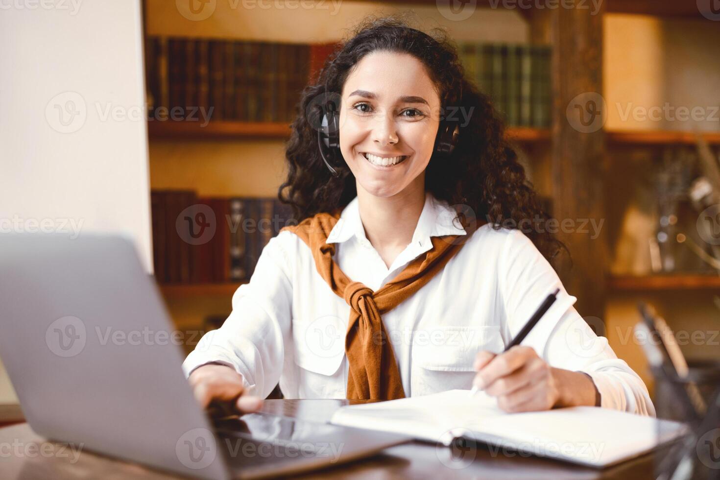 Remote Education During Quarantine. Portrait of cheerful woman sitting at table with pen, writing in notebook, using laptop and taking notes. Positive female student learning, looking at camera photo