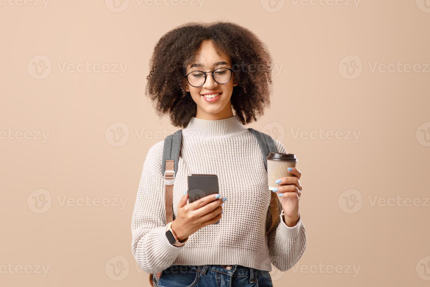 Modern gadgets for communication remotely. Smiling african american woman in glasses with backpack holds cup of coffee and typing on smartphone, isolated on yellow background, studio shot, copy space photo