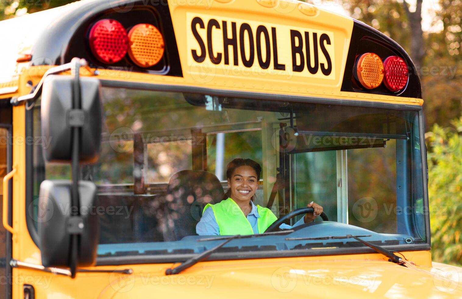 Portrait Of Happy Black Female Driver Driving Yellow School Bus, Beautiful Young African American Lady In Reflecting Vest Looking Through Window, Holding Steering Wheel An Smiling At Camera photo