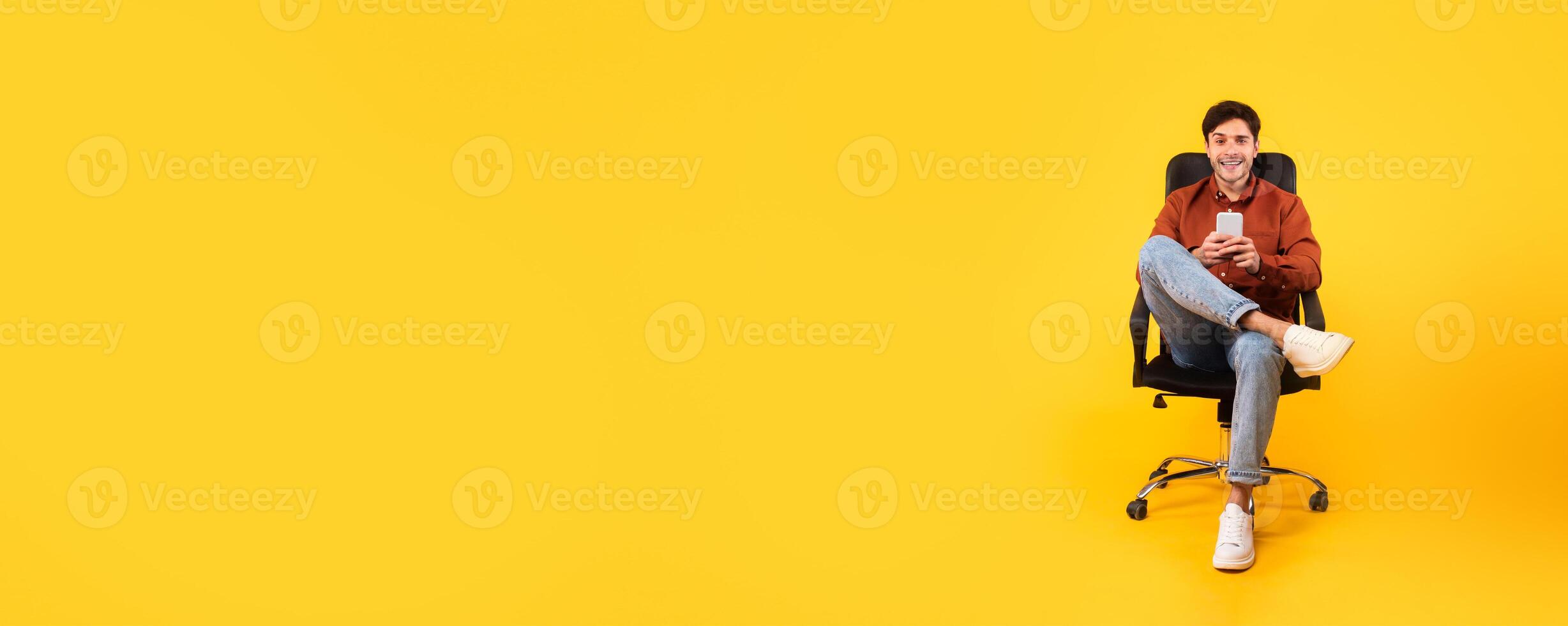 A young man relaxes in a black office chair, holding a drink. He is dressed casually, with a cheerful expression against a vivid yellow background, giving a lively and inviting atmosphere. photo