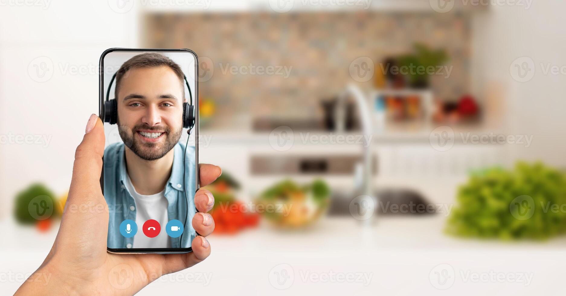 A person holds a smartphone showing a smiling individual on a call. The background features a cozy kitchen with fresh vegetables, highlighting a casual conversation atmosphere. photo