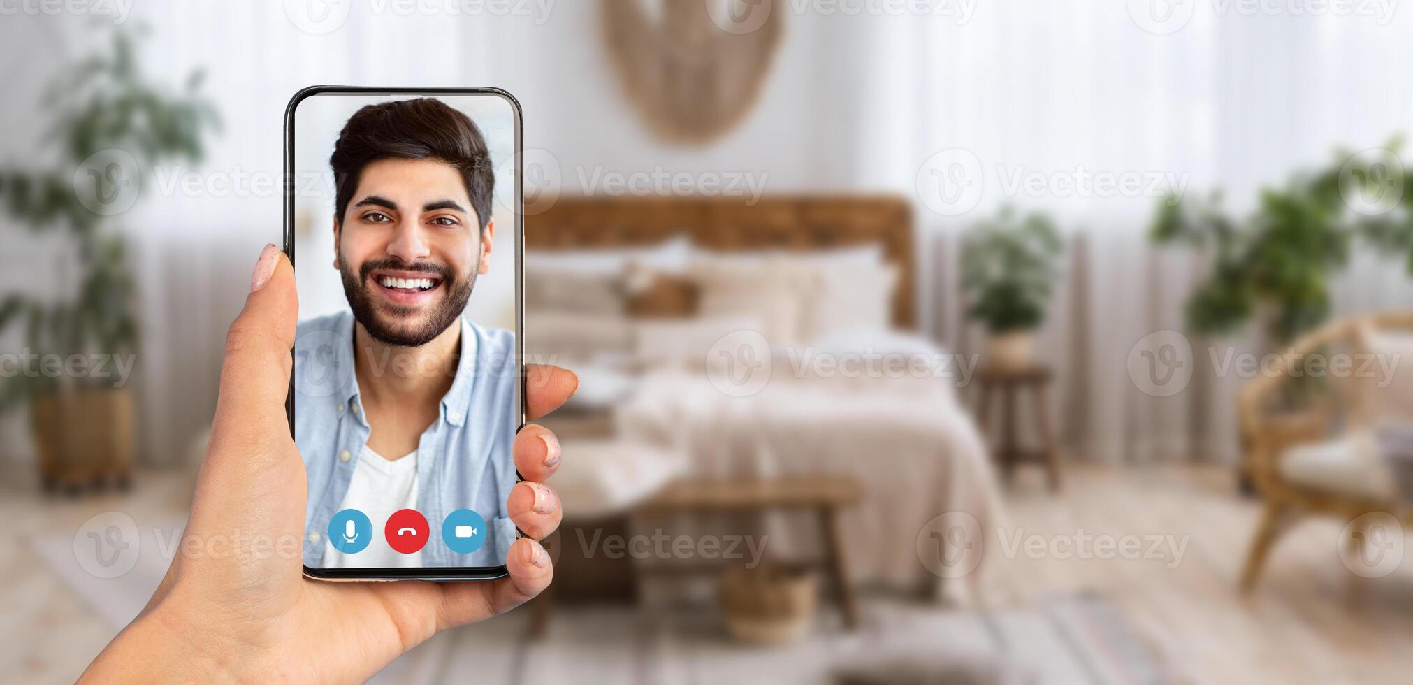 A person holds a smartphone showing a smiling face on a call. The background reveals a comfortable room decorated with plants and soft furnishings, creating a warm atmosphere for communication. photo
