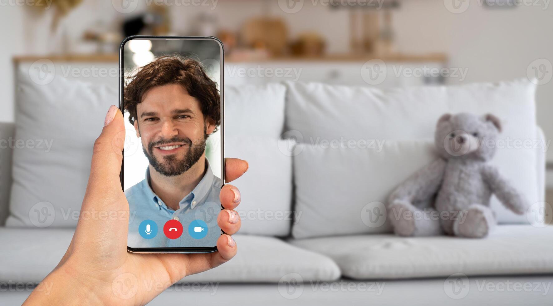 A person participates in a call, holding a smartphone with a smiling face on the screen. The setting is a cozy living room with a couch and a teddy bear, creating a warm atmosphere. photo