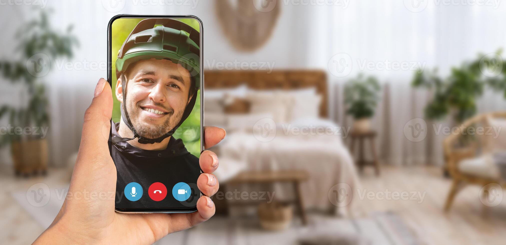 A person smiling while on a call appears on a smartphone screen. The background features a well-decorated home interior with light curtains and comfortable furniture. photo