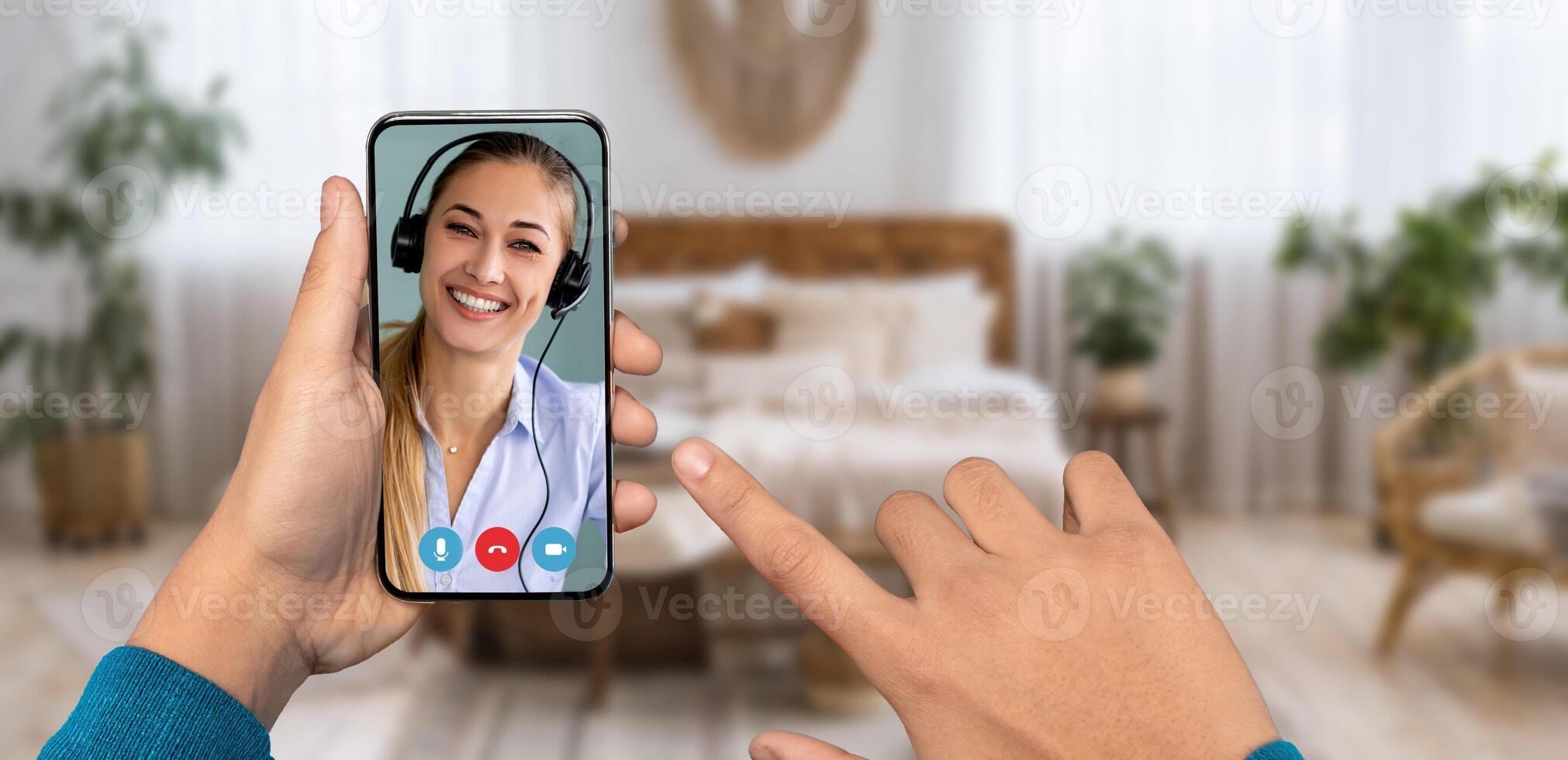 A person is using a smartphone to participate in a call, interacting with a smiling individual wearing a headset. The setting is a cozy home environment with soft furnishings. photo