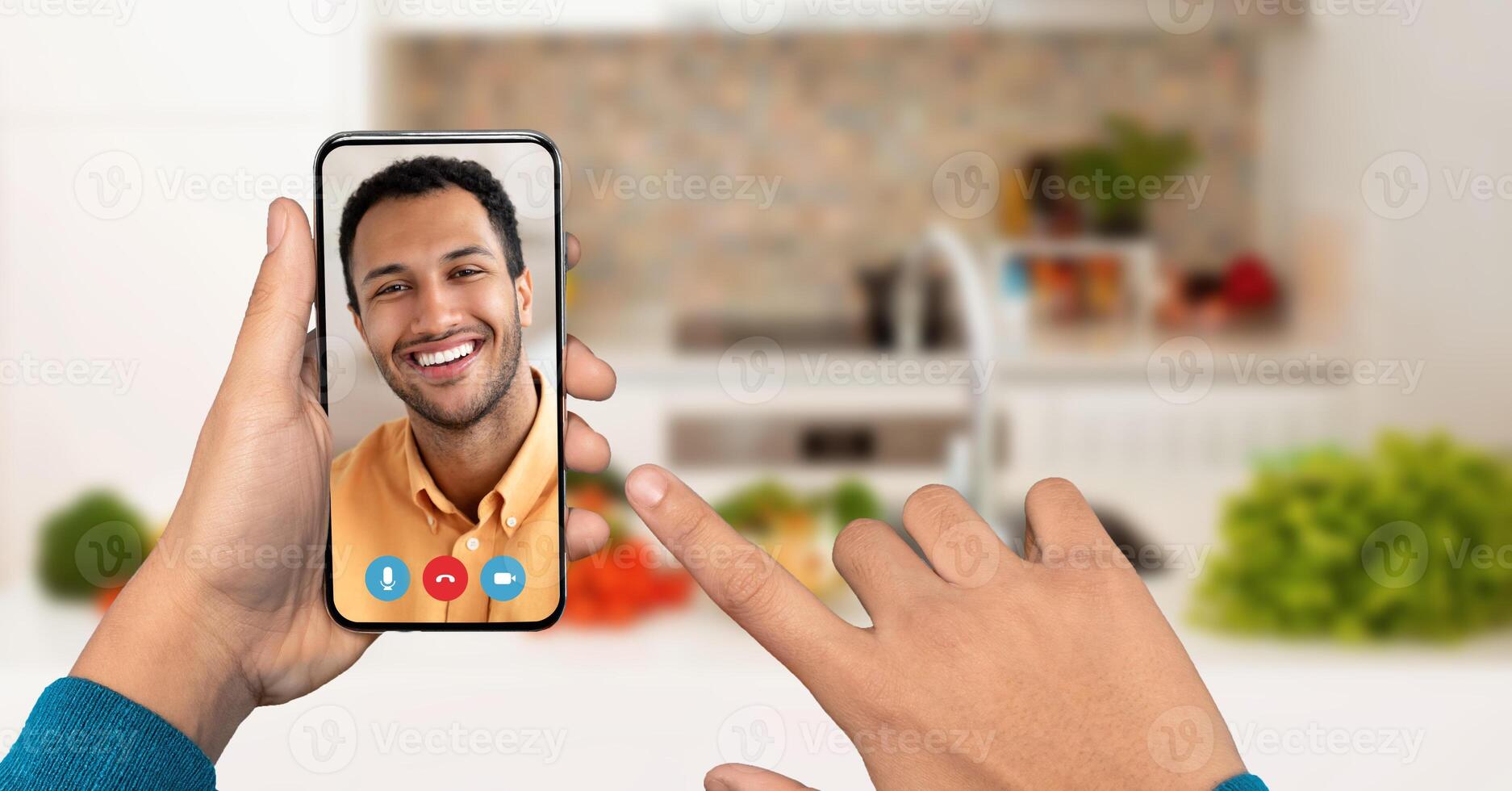 A person engages in a call using a smartphone in a bright kitchen, pointing at the screen with a smiling face of another individual visible. Fresh vegetables are placed in the background. photo