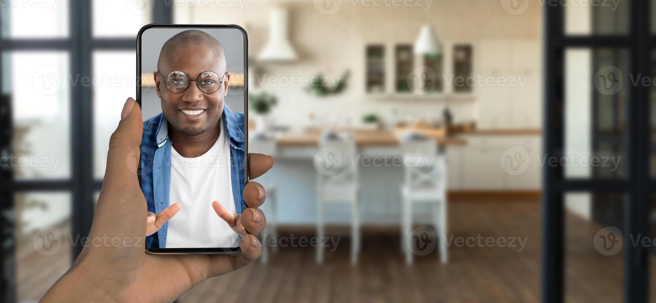 A person holds a smartphone displaying a smiling individual during a call. The setting features a bright kitchen with wooden floors and a modern, inviting atmosphere. photo