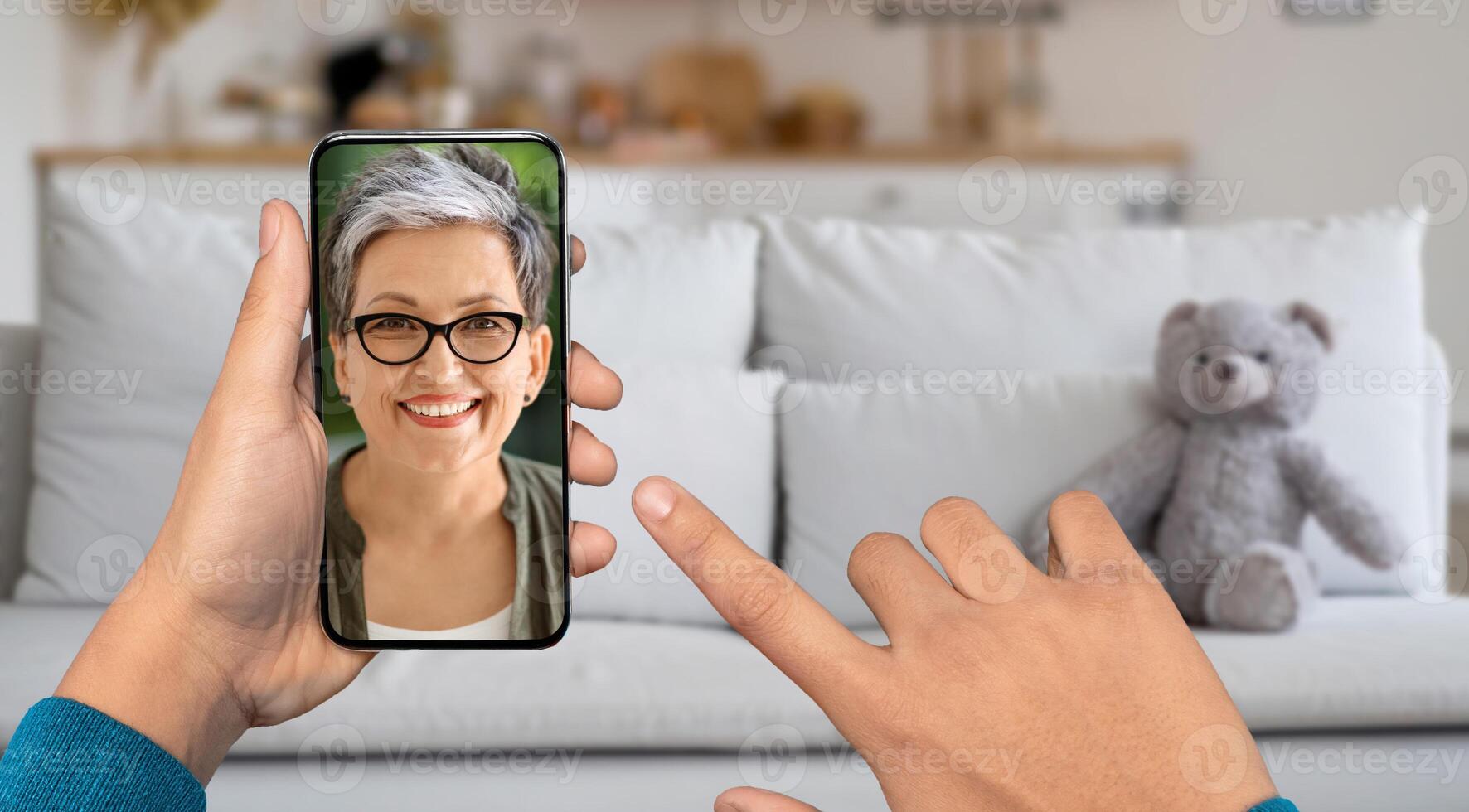 A person is using a smartphone for a call from their home. The lady on screen smiling and engaged in conversation while relaxing on a couch with a teddy bear nearby. photo